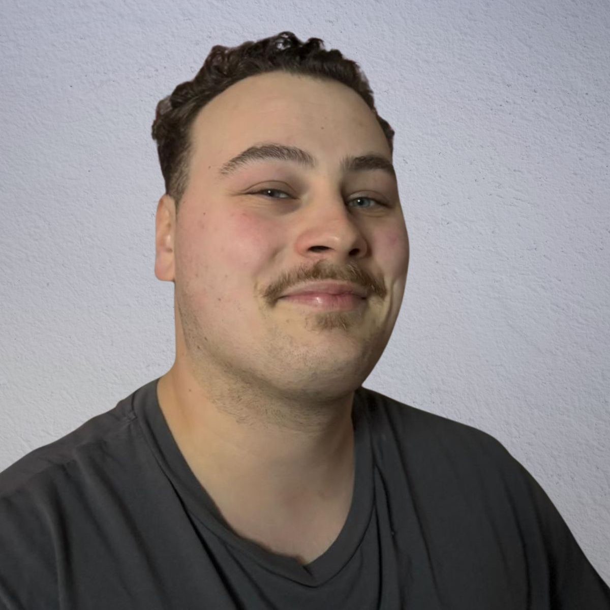 Ein junges Mann lächelt mit einem selbstbewussten Ausdruck, trägt ein graues T-Shirt.