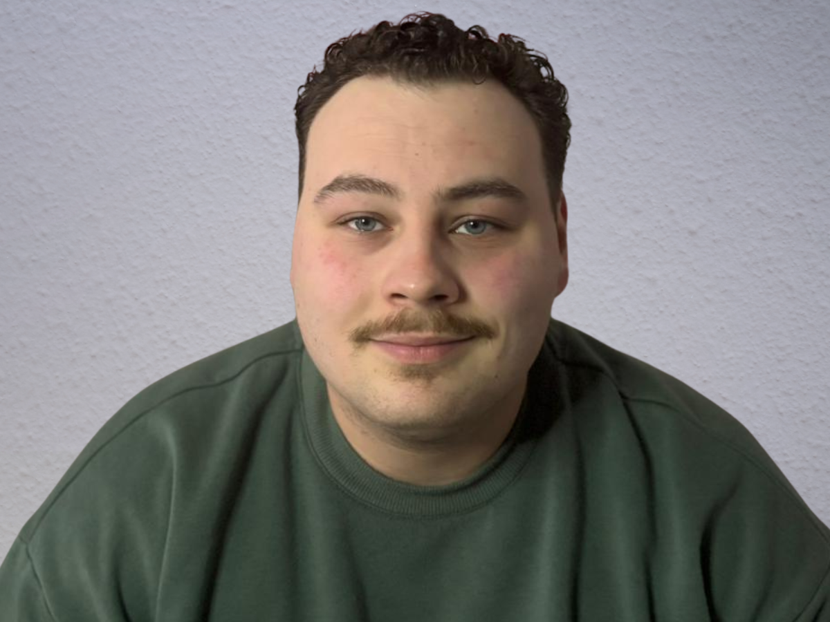 Junger Mann mit lockigem Haar und Schnurrbart, der in die Kamera lächelt.